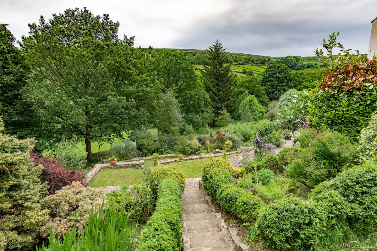 A terraced garden with stone steps leading down to a lush landscape of various trees and flowering shrubs. Rolling hills can be seen in the background, enhancing the natural beauty of the countryside setting. The garden offers a peaceful retreat surrounded by greenery.