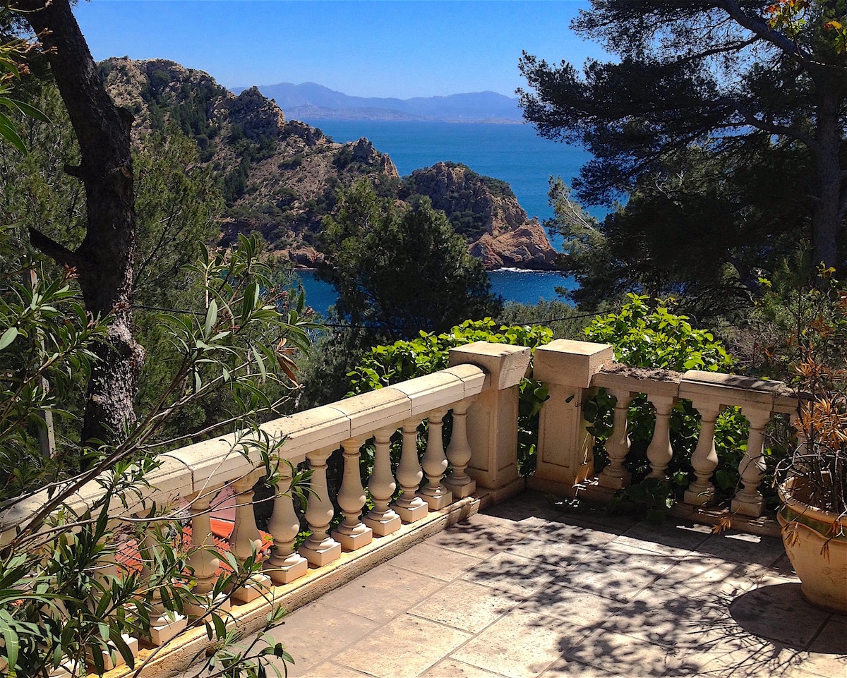 A terrace adorned with a decorative stone railing overlooks a tranquil sea and rocky coastline. Lush greenery and terracotta pots are present, with the sunlight casting gentle shadows across the tiled floor. The distant mountains are visible under a clear blue sky.