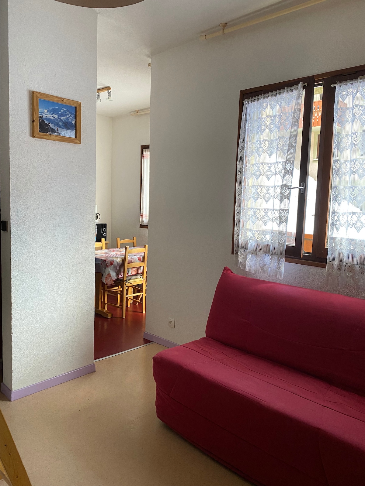 A bright living area features a pink sofa, positioned against a white wall. Natural light filters through a lace curtain covering a window. An archway leads to a dining area with wooden chairs and a table, visible in the background.