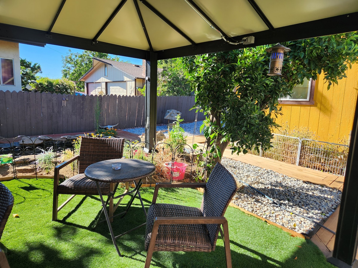 An outdoor seating area features a round table accompanied by two chairs, all set on a grassy surface. Surrounding plants include shrubs and a boundary of wooden fencing, providing a serene space for relaxation. A few gardening supplies are visible in the background.
