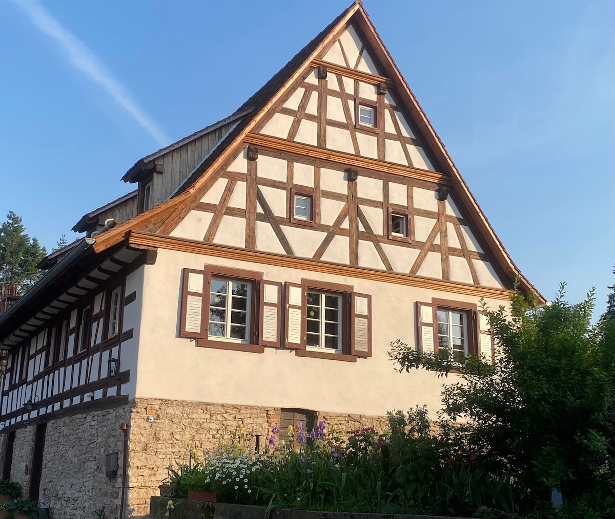 A traditional half-timbered house is depicted, featuring a stone and wood exterior. The multi-pane windows are adorned with shutters, allowing natural light to enter. A vibrant flowerbed fills the foreground, adding color to the rustic charm of the structure.