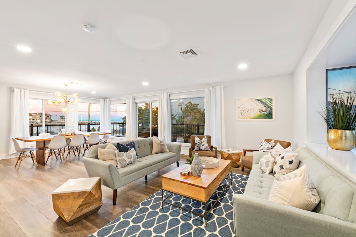 A bright and spacious living area showcases modern furniture with comfortable sofas and decorative pillows. Large windows allow natural light and offer views of the ocean. A stylish dining table with seating for eight complements the refined decor, enhancing the inviting atmosphere.