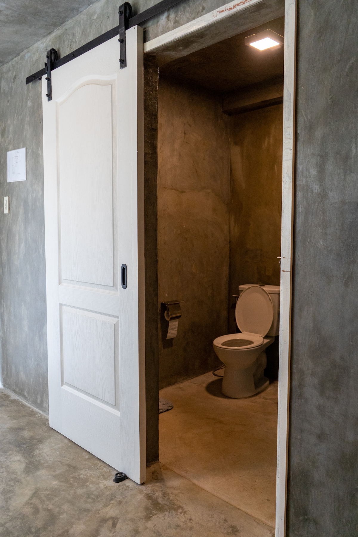 An interior space features a sliding white door leading to a bathroom. The bathroom presents a simple design, showcasing a white toilet against a textured grey wall. The floor is a neutral tone, complementing the modern industrial style.