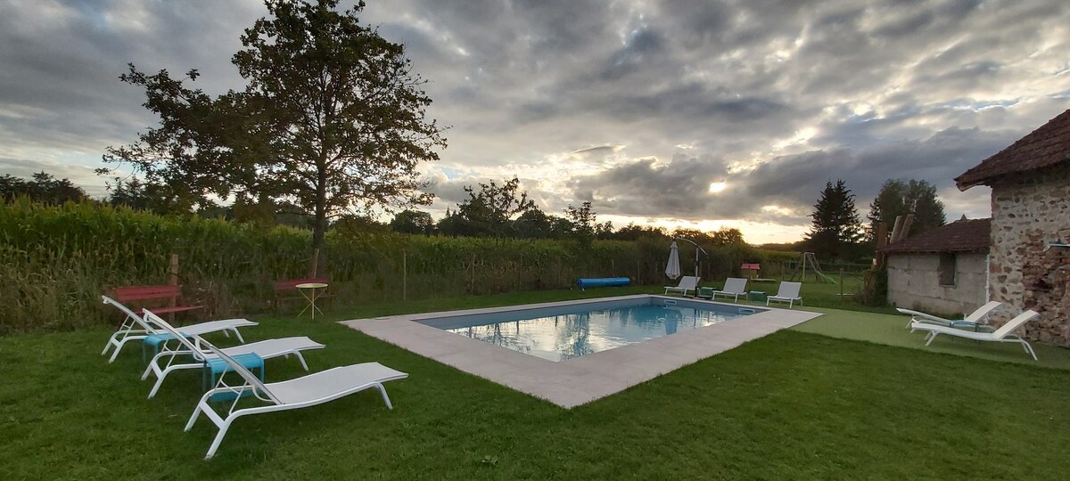 An outdoor space features a heated swimming pool surrounded by a grassy area. Lounge chairs are arranged nearby, complemented by tables positioned among the greenery. The backdrop includes a cornfield and a cloudy sky, creating a serene environment.