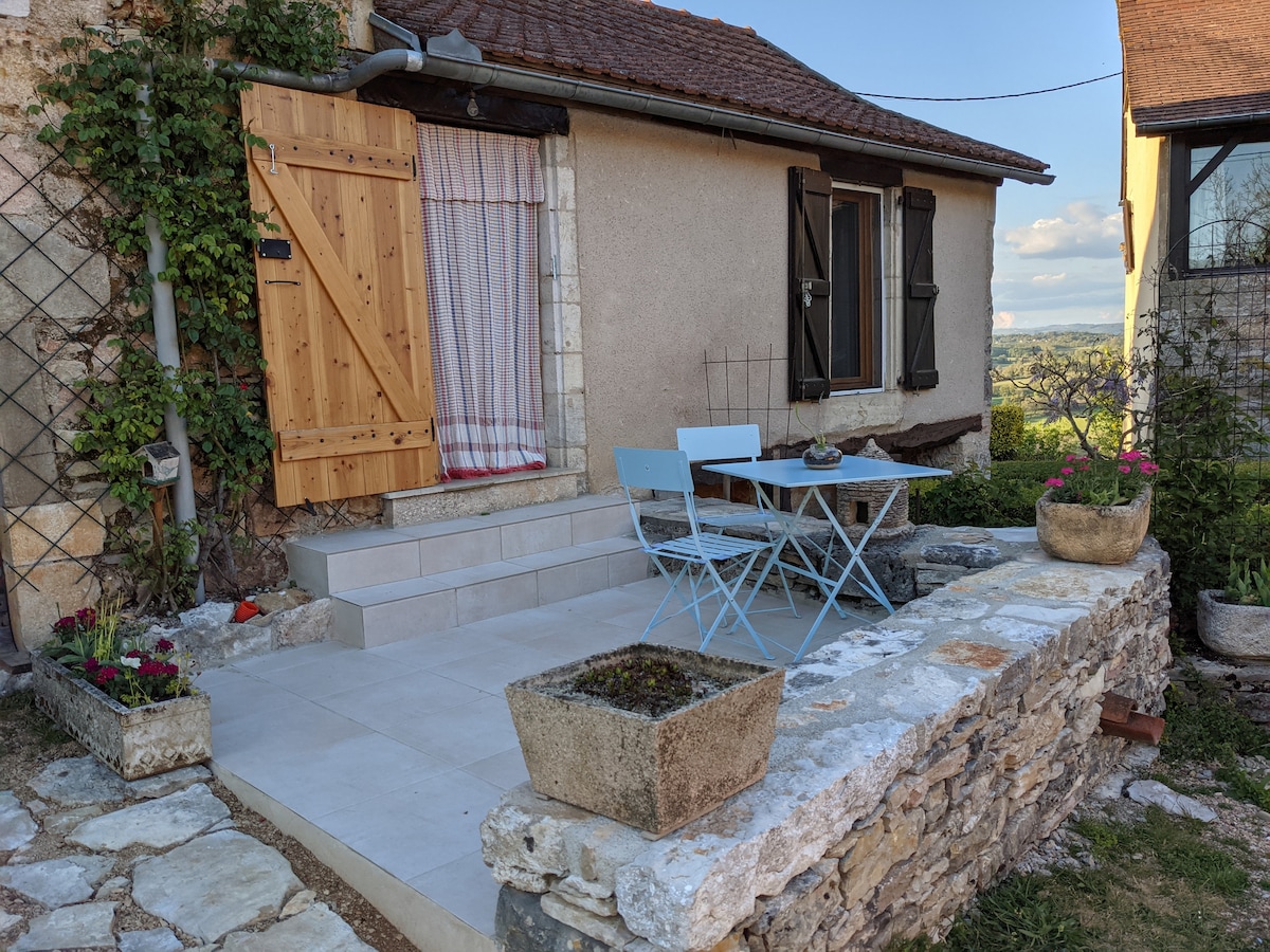 A small terrace features a light blue table with two matching chairs, surrounded by potted plants. The entryway is adorned with a wooden door and a partially opened curtain, while the stone steps lead up to the door of the studio.