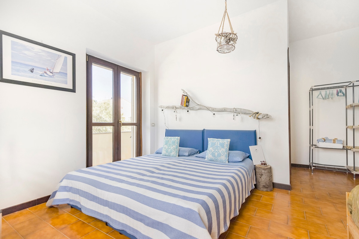 A spacious room features a large striped bedspread in blue and white, complemented by two decorative pillows. Natural light enters through two large windows, framed by a simple decor. A decorative shelf made from driftwood adds a coastal touch, enhancing the serene ambiance.