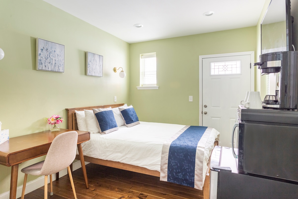 A well-lit private room is presented with green walls and natural light from the door and windows. A spacious bed with blue and white linens sits beside a work desk and a mini fridge. Decorative wall art adds subtle charm to the space.