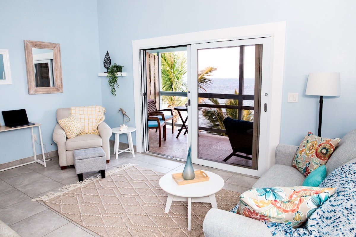 A bright living space is furnished with a light gray sofa and a cozy armchair. A small desk with a laptop is positioned nearby. Sliding glass doors reveal a view of the ocean and outdoor seating, framed by palm trees.