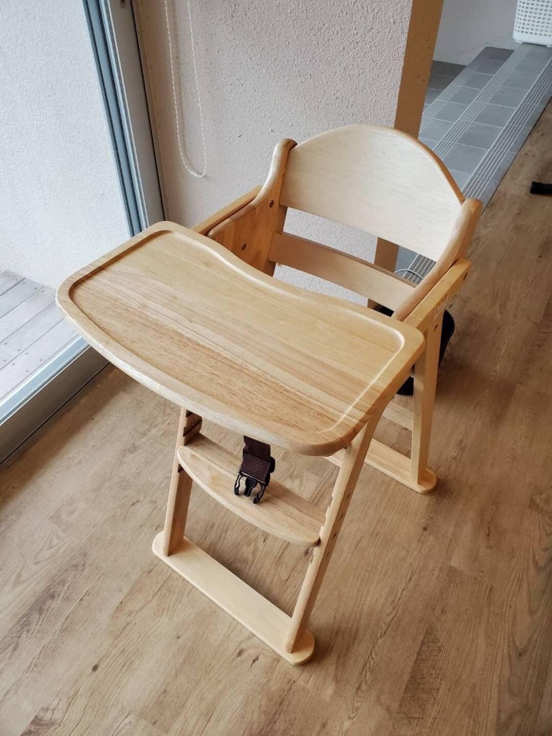 A wooden high chair is positioned against a wall, featuring a smooth, well-polished surface. The chair includes a large, flat tray and safety straps for secure seating, with a minimalist design and light wood color that complements the surrounding space.