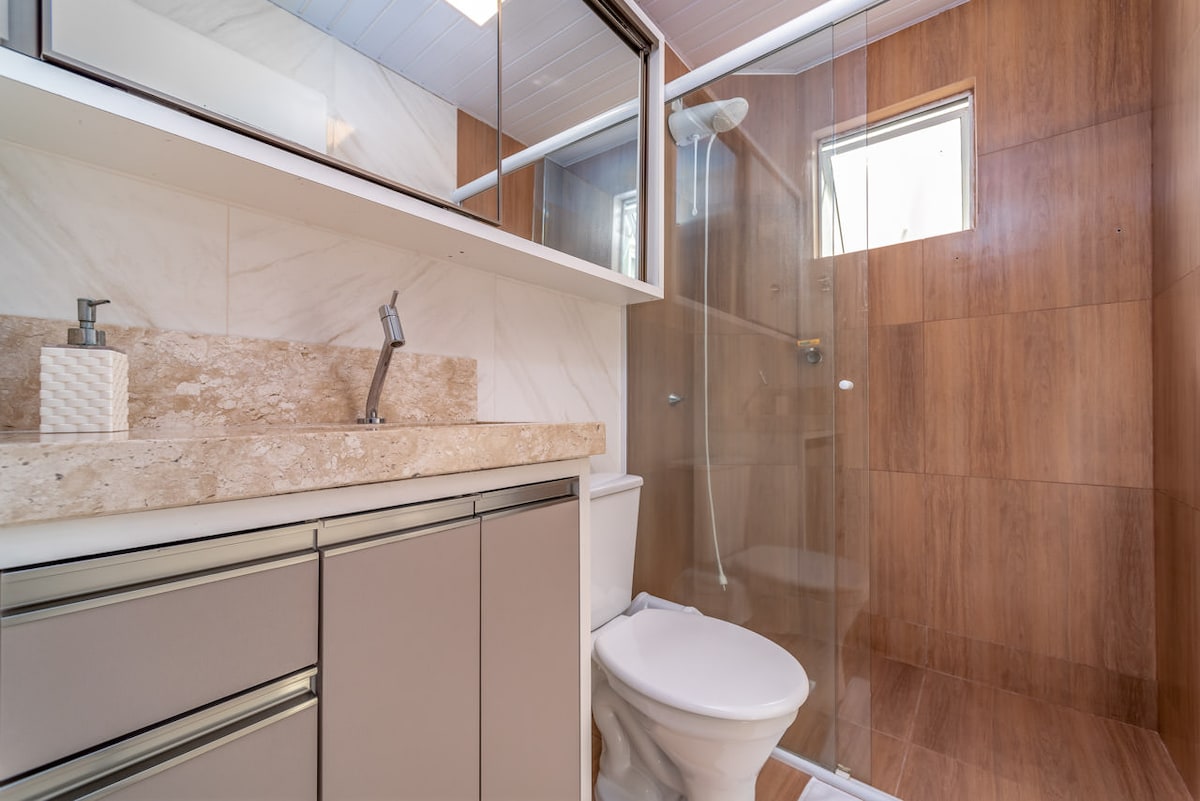 A modern bathroom features a spacious shower enclosed in clear glass. A sleek vanity with a light marble countertop is paired with neutral cabinetry. Natural light enters through a small window, which complements the warm wood-like tiles.