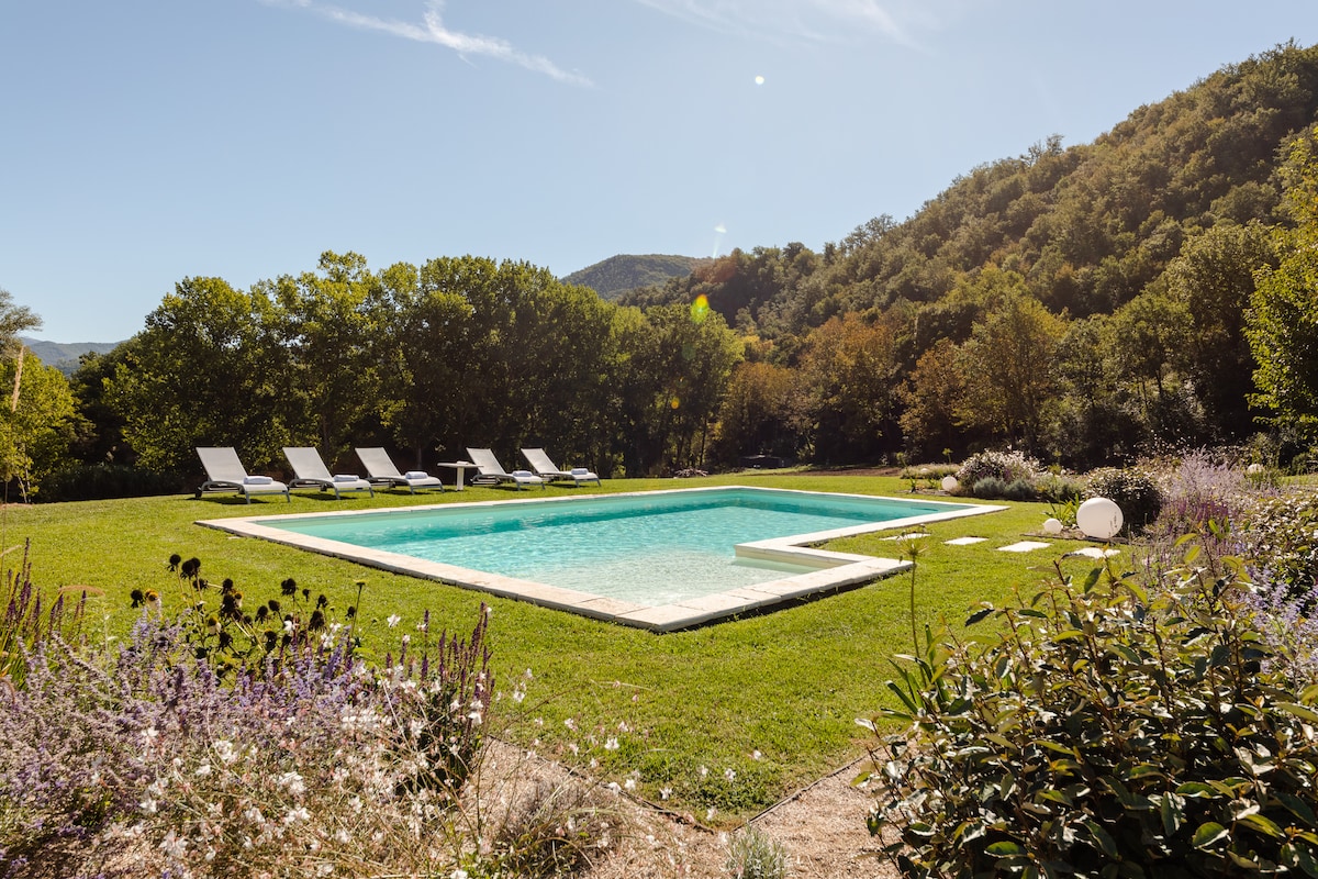 A spacious outdoor swimming pool is surrounded by well-maintained greenery and blooming flowers. Sun loungers are positioned nearby, inviting relaxation in the sun. Lush trees and rolling hills provide a serene backdrop, enhancing the peaceful ambiance of this private oasis.