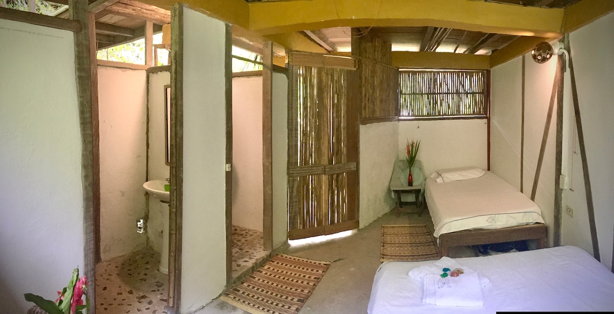 The room features a spacious layout with natural materials, including bamboo accents. Two sleeping areas are present, one with a cozy bed and the other with an additional sleeping space. A private bathroom area is separated by a wooden partition, adding to the room's openness.