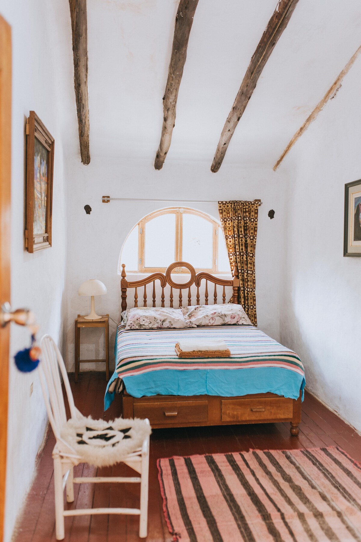 A bright sleeping area features a wooden bed with a striped blanket laid across it. An arched window allows natural light to fill the room, complemented by decorative curtains. A wooden chair and a small lamp are positioned nearby, enhancing the simplicity of the space.