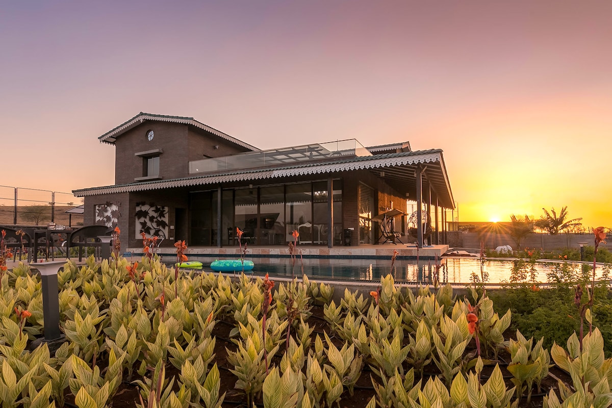 The villa is showcased at sunset, emphasizing its modern design with large glass windows and a flat roof. Surrounding lush green plants frame the property. A serene swimming pool reflects the warm hues of the sky, creating a peaceful outdoor setting.