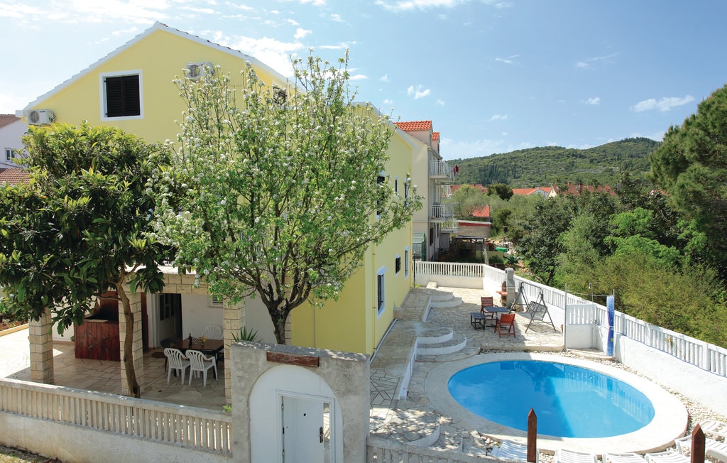 The exterior of a yellow building is shown, surrounded by greenery. A circular swimming pool is visible, with a sunbathing area featuring loungers. An outdoor patio with tables and chairs is seen, along with trees providing shade in the vicinity.