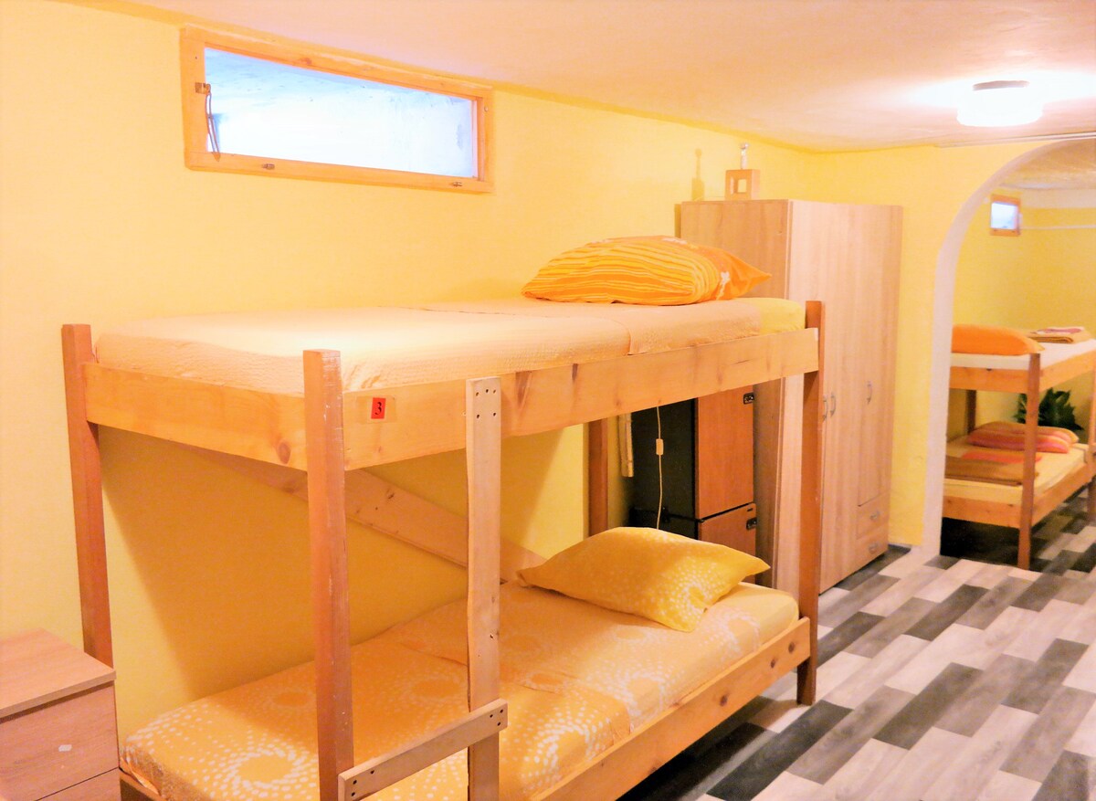 A mixed dormitory room features a wooden bunk bed with a bottom single bed and bright yellow bedding. A small nightstand is beside the beds. A wardrobe stands against the wall, while narrow windows provide natural light to the space.