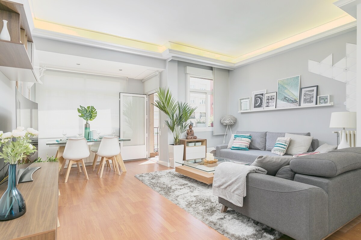 A light-filled living area features a cozy gray sofa adorned with decorative cushions. A wooden table is set for dining, while a large plant adds a touch of greenery. Floor-to-ceiling glass doors lead to a terrace, enhancing the open and airy feel.