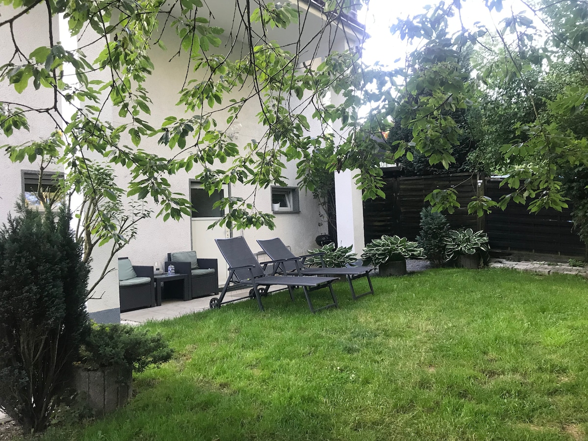 A well-maintained garden area features green grass and a few lounge chairs positioned for relaxation. Potted plants are placed along the edges, while a covered seating area offers shaded comfort. Trees provide additional natural surroundings, enhancing the peaceful atmosphere of the outdoor space.