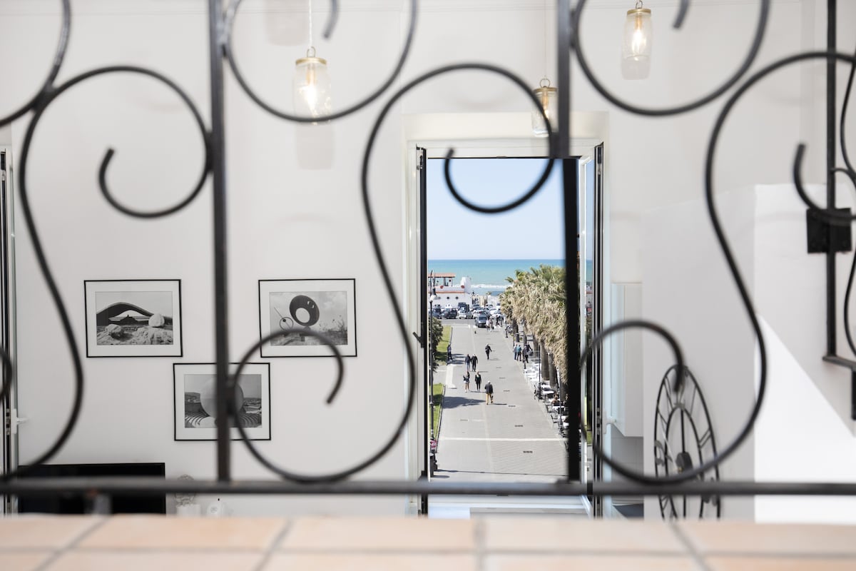 A view from a balcony features a scenic coastal promenade lined with palm trees. The ocean is visible in the distance, inviting relaxation. Artistic black-and-white photographs adorn the walls, enhancing the space's character. Natural light streams through the open doors, creating a bright atmosphere.