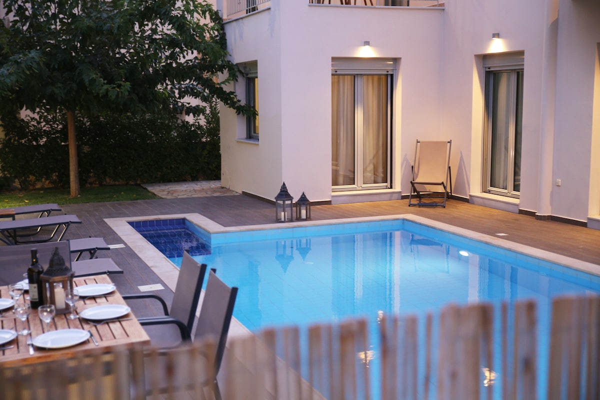 A private swimming pool is surrounded by a wooden deck and lounge chairs. Elegant lanterns add a subtle ambiance. The villa's architecture features large windows that reflect natural light, while lush greenery provides a natural border, enhancing the tranquil outdoor oasis.