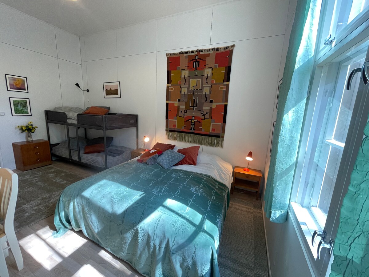 A spacious room features two single beds in a metal frame, and a large double bed covered with an aqua-colored blanket. A decorative wall hanging adds color, and natural light streams in through large windows, illuminating the simple furnishings and inviting atmosphere.