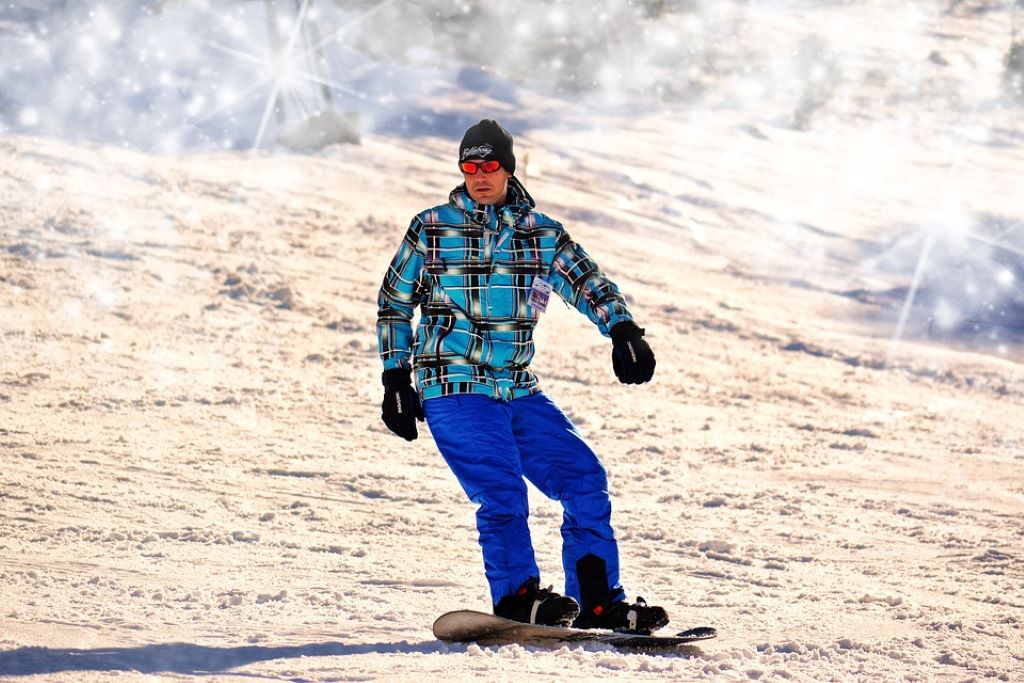 A snowboarder is shown carving through fresh snow on a mountain slope. The individual is dressed in a patterned blue jacket and blue pants, with snowy terrain and a light flurry of snowflakes in the background adding to the winter atmosphere.