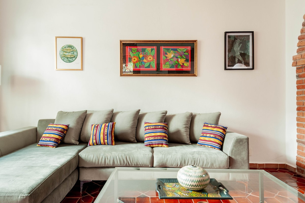 A spacious living area is featured, showcasing a light-colored sectional sofa adorned with vibrant, patterned throw pillows. A glass coffee table sits in front, while artwork is displayed on the walls, adding color to the bright and airy space.