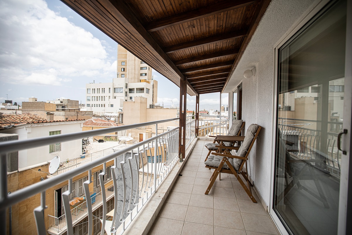 A covered balcony extends the living space, showcasing a mix of urban architecture. Two wooden lounge chairs are placed beside a glass railing, offering unobstructed city views. Natural light filters through, highlighting the tiled flooring.