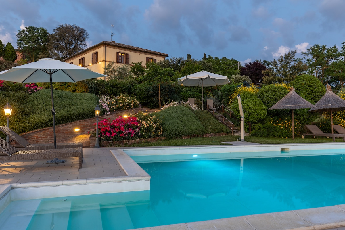 A serene swimming pool reflects the evening sky, surrounded by lush greenery and vibrant flower beds. Umbrellas provide shade over lounge areas, enhancing the tranquil outdoor space. The historic casale is visible in the background, blending with the natural landscape.