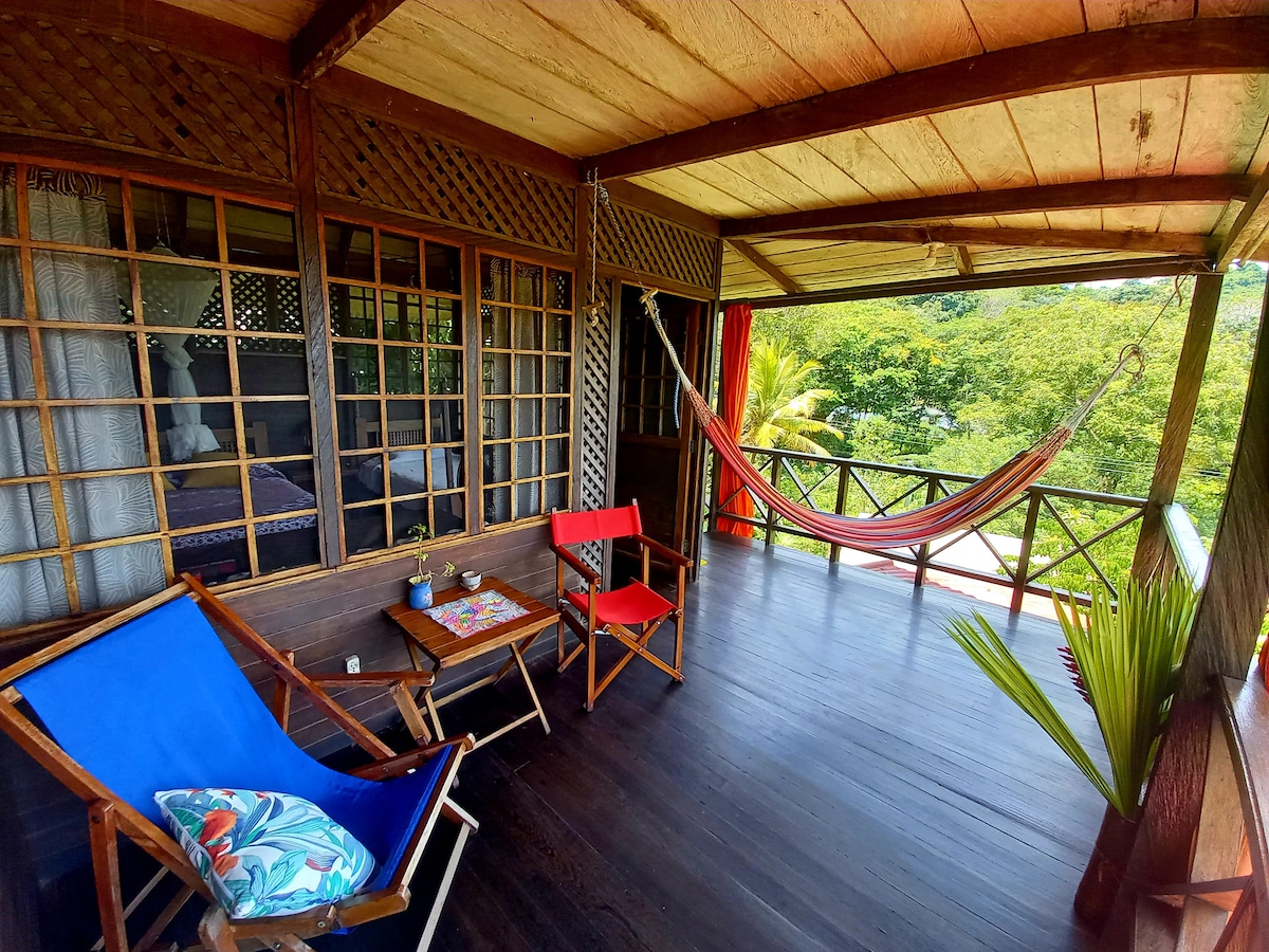 A shaded wooden porch features a hammock strung between posts, with a vibrant blue chair and a red chair positioned nearby. A small table holds decorative items, while the surrounding greenery offers a serene view of the landscape.