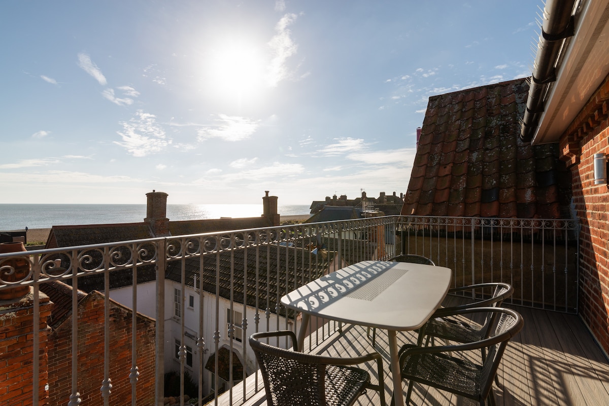 A balcony furnished with a table and four chairs overlooks the sea and surrounding rooftops. The bright sky is partly cloudy, with sunlight illuminating the scene. The railing offers safety while enjoying the expansive coastal views.