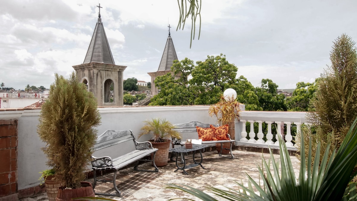 A spacious terrace is adorned with two metal benches and potted plants, providing a comfortable area to relax. The view includes two distinct church towers framed by lush greenery, inviting a connection to the surrounding historic architecture.