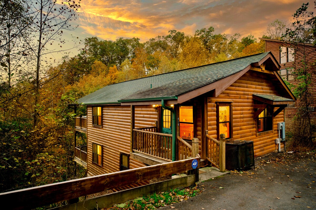 A charming log cabin is nestled among vibrant autumn foliage, illuminated by the warm glow from its windows. The exterior showcases a wooden deck, enhancing the inviting appearance of the home. The surrounding landscape features colorful leaves from nearby trees, blending beautifully with the rustic atmosphere.