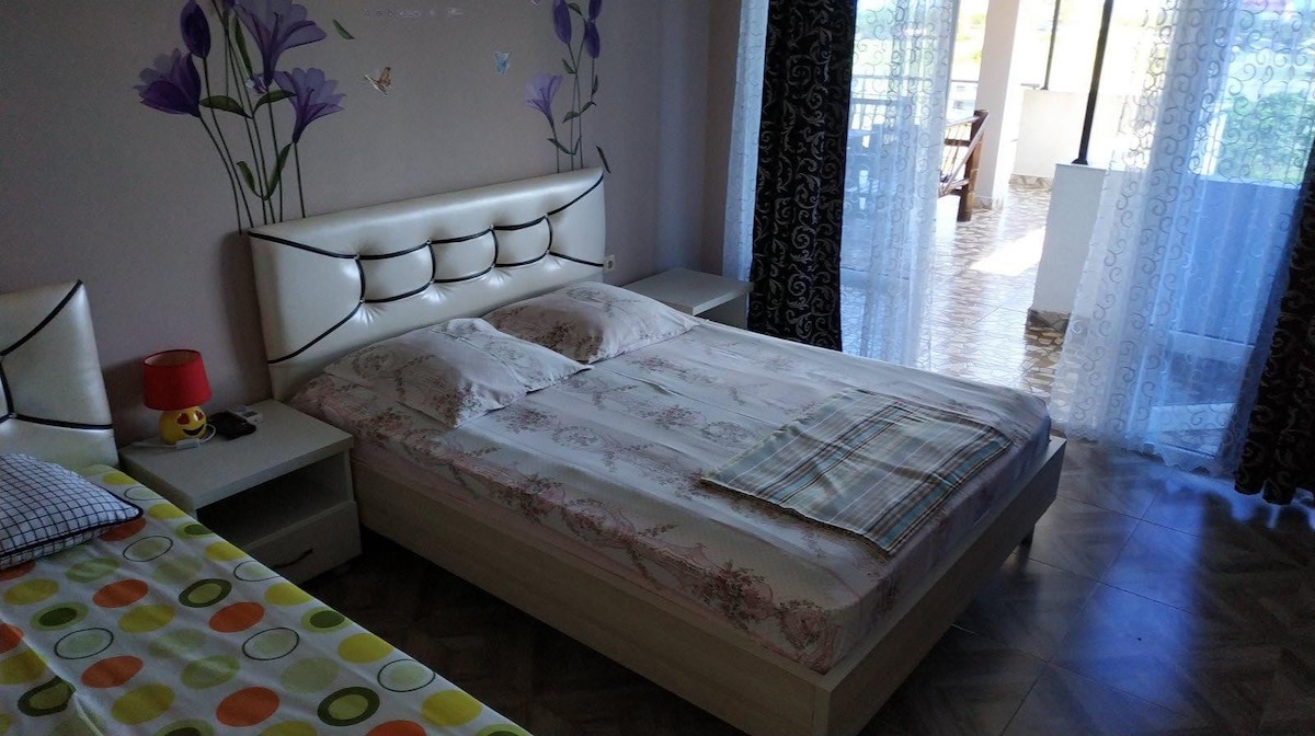A spacious sleeping area features a light-colored bed with patterned bedding. A small bedside table with a lamp is positioned nearby. Large windows allow natural light to fill the room, which is adorned with wall art showcasing floral designs.