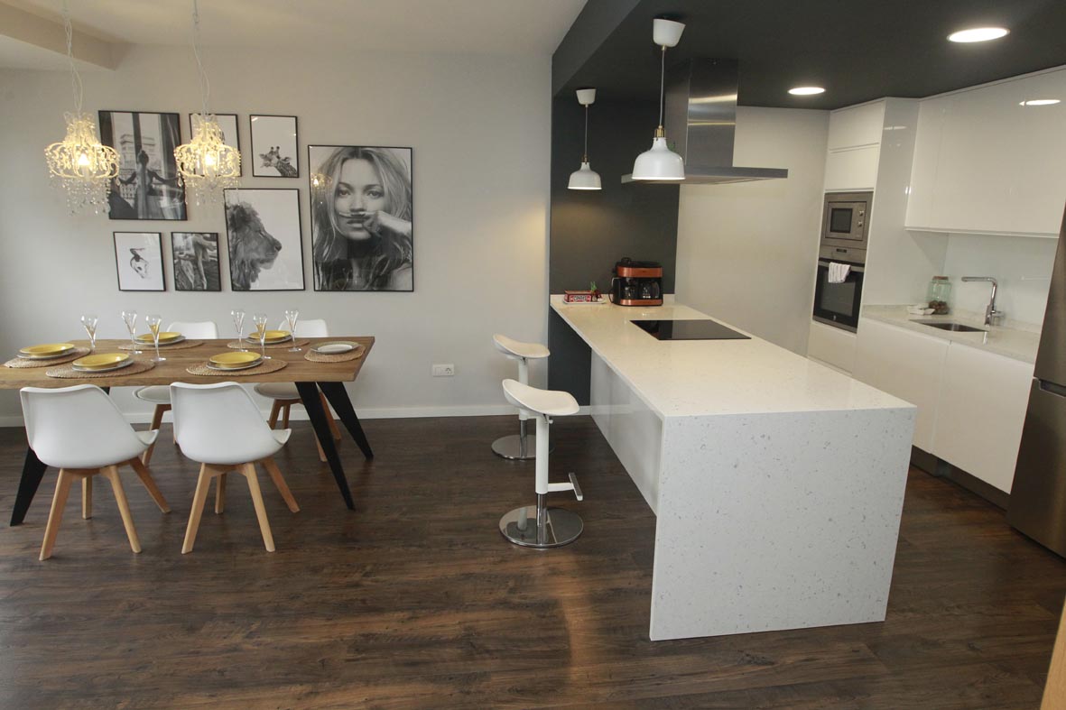 A modern kitchen and dining area feature sleek white cabinetry and a spacious island with a built-in stovetop. A dining table set for six is complemented by simple, stylish chairs. Photos decorate the wall, enhancing the contemporary atmosphere of the space.