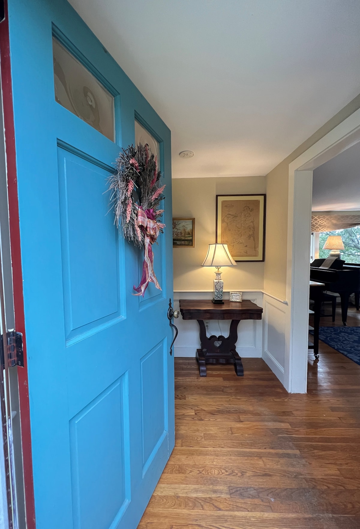 A brightly colored blue door welcomes guests, adorned with a seasonal wreath. Inside, a warmly lit entryway reveals a small table with a lamp, providing a cozy introduction to the space. The living area is visible through an adjacent doorway.