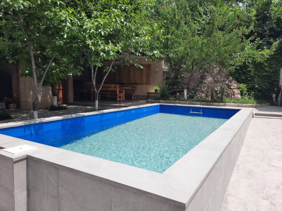 A clear blue swimming pool is surrounded by lush greenery and several trees. Stone pathways lead to seating areas in the background, where dining and relaxation spaces are visible. The pool is designed with a modern aesthetic, featuring clean lines and a smooth edge.