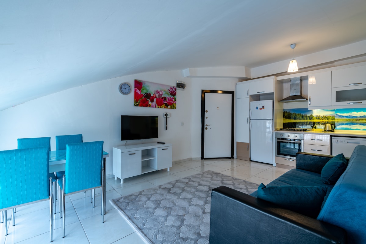 A modern apartment interior is depicted, featuring a blue sofa and a dining area with four turquoise chairs. A flat-screen TV is mounted on a white cabinet, and a fully equipped kitchen is visible with stainless steel appliances. Colorful artwork adorns the wall.