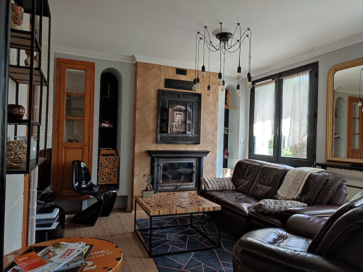 A comfortable living area features a dark leather sectional sofa facing a textured fireplace. A large coffee table with a unique design sits at the center, complemented by a black chair. Natural light pours in through the window, enhancing the space's warmth.