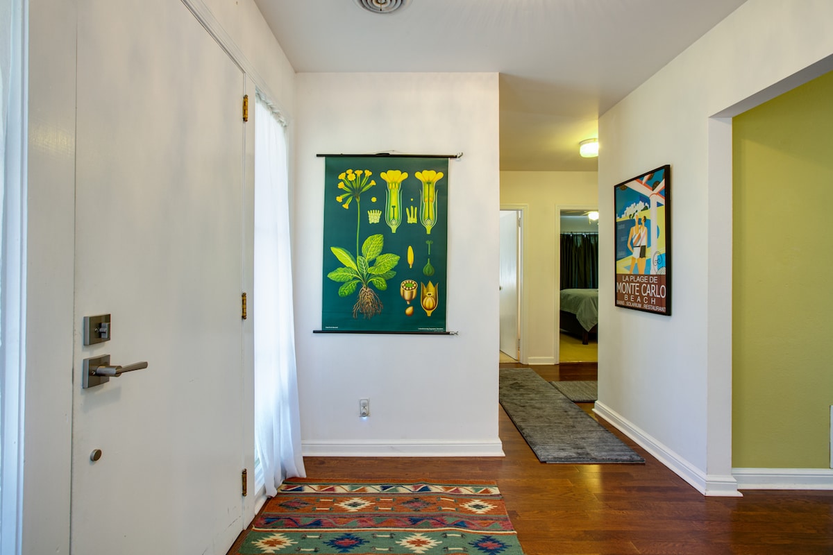 A welcoming entrance area features hardwood flooring and a colorful area rug. A botanical wall hanging adds visual interest, while light enters through a nearby window. The hallway leads to additional spaces in the home, indicating a well-connected layout.
