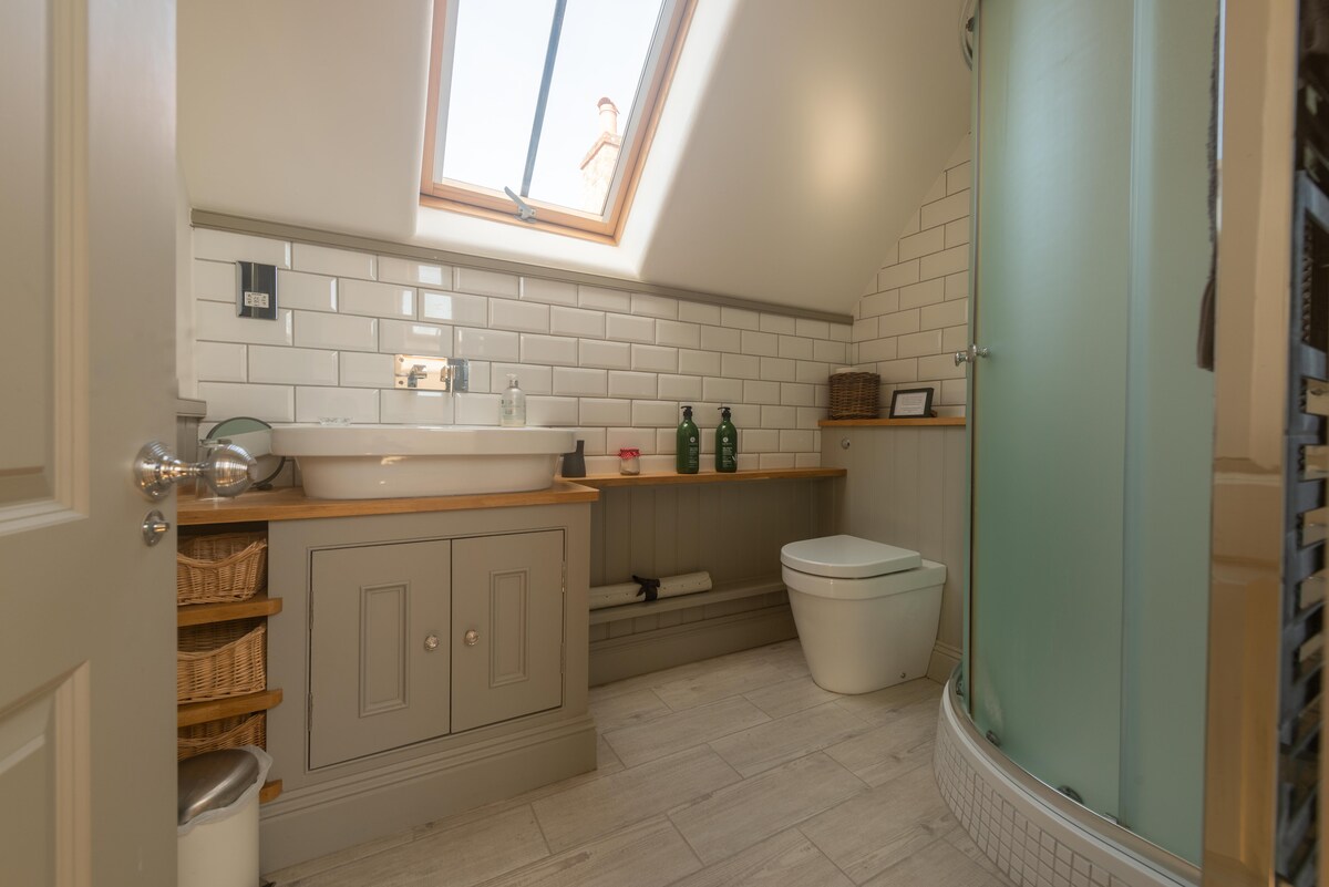 A modern bathroom displays a spacious sink atop a wooden counter, with green bottles arranged neatly. A glass shower enclosure stands adjacent, and natural light filters in through the skylight, illuminating the white tiled walls and light wood flooring, creating an airy atmosphere.