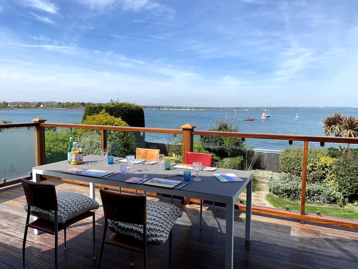 An outdoor dining table is set on the large deck, overlooking scenic views of the water and distant boats. Colorful chairs surround the table, which is adorned with tableware and a refreshing drink. Lush greenery and well-kept gardens are visible in the background.