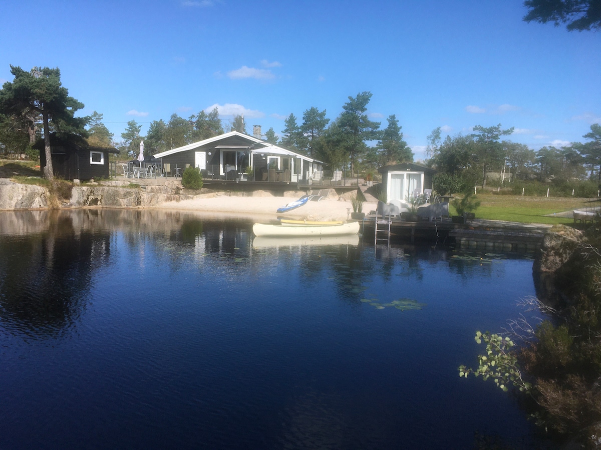 A tranquil scene is presented, featuring a calm body of water with a canoe resting atop its surface. In the background, a spacious cabin is surrounded by trees, and a private sandy beach is visible, enhancing the serene outdoor setting.