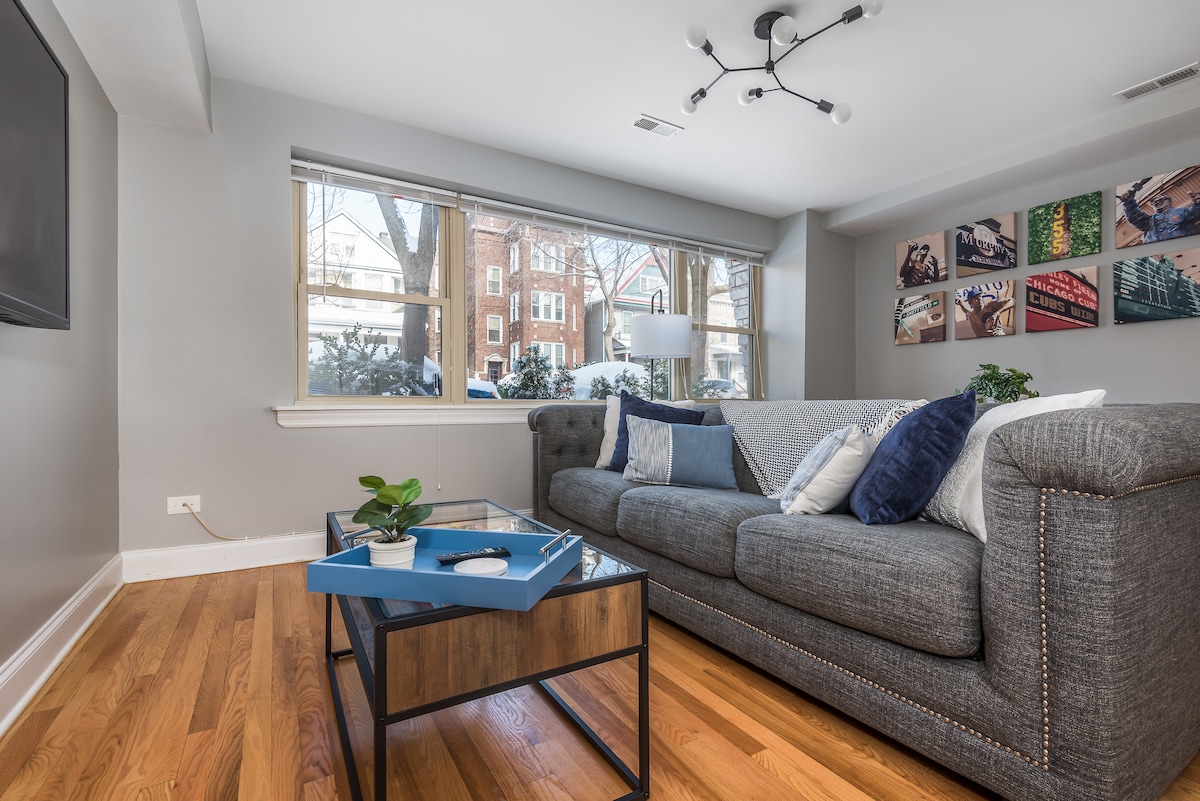 An inviting living area features a spacious gray couch adorned with blue and white cushions. A modern coffee table with a decorative tray sits on polished hardwood floors, while large windows allow natural light to fill the space, providing views of the surrounding greenery.