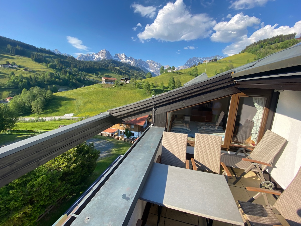 An elevated terrace showcases outdoor furniture, including a table and four chairs. A panoramic view of lush green hills and snow-capped mountains is visible in the background, enhancing the sense of open space and natural beauty.