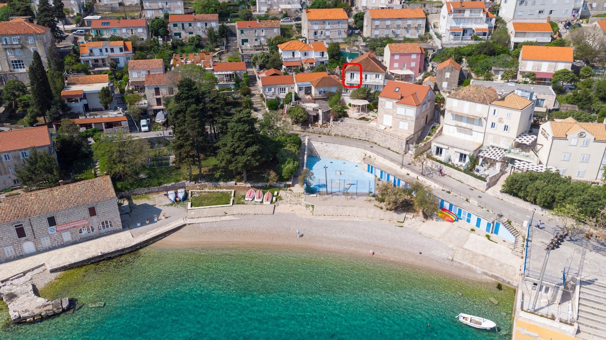 An aerial view captures a quaint coastal neighborhood, showing a mix of houses with red rooftops. The clear turquoise water and sandy beach are visible at the foreground, with a gentle curve of the shoreline inviting visitors to explore. A few boats are anchored near the beach.