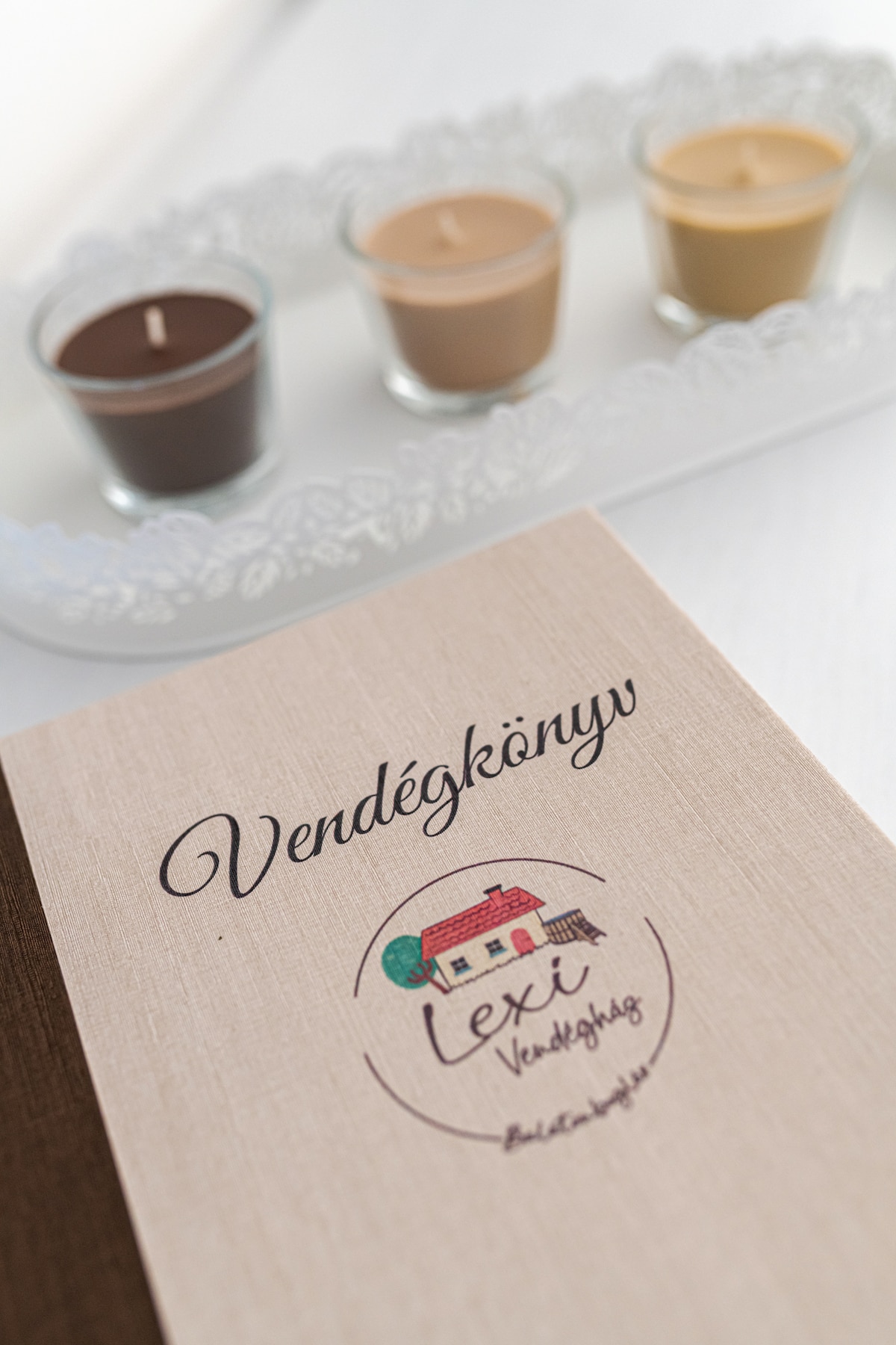 A guest book titled 'Vendégkönyv' is placed on a flat surface. The design includes a simple illustration of a house, accompanied by three small candles in varying shades of brown, displayed on a decorative tray.