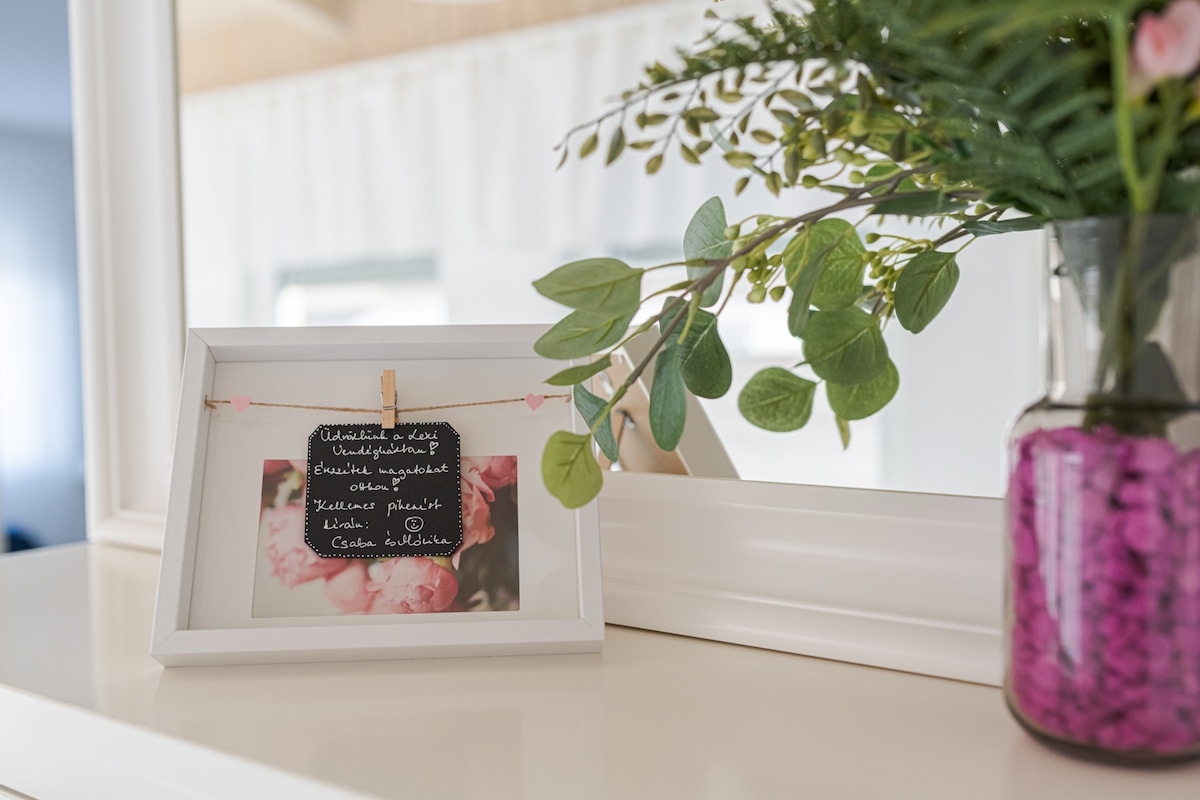 A decorative frame containing a floral image is displayed on a white surface. A small chalkboard sign hangs from twine, while a glass vase filled with purple stones holds fresh greenery, enhancing the serene aesthetic of the area.