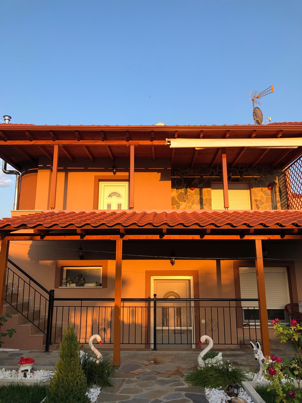 A two-story villa with an orange hue features a terracotta roof and a welcoming front porch. Decorative elements, including swans and a well-maintained garden, enhance the exterior. The stairway leads to the entrance, framed by windows that provide a glimpse of the interior.