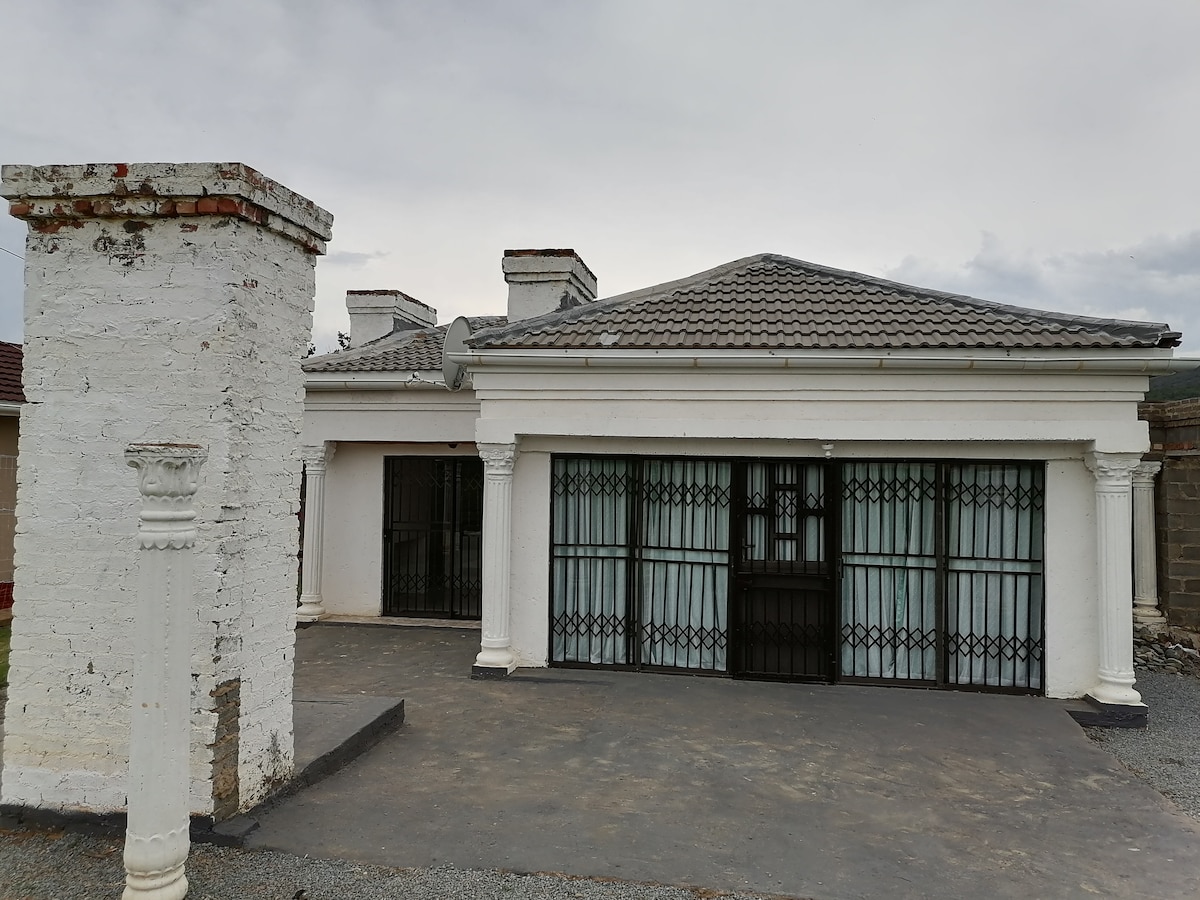A single-story building with a symmetrical design is featured, showcasing a sloped rooftop and decorative white columns. The entrance includes a gated area with large windows, allowing natural light to filter inside. A clean, paved pathway leads to the front door.